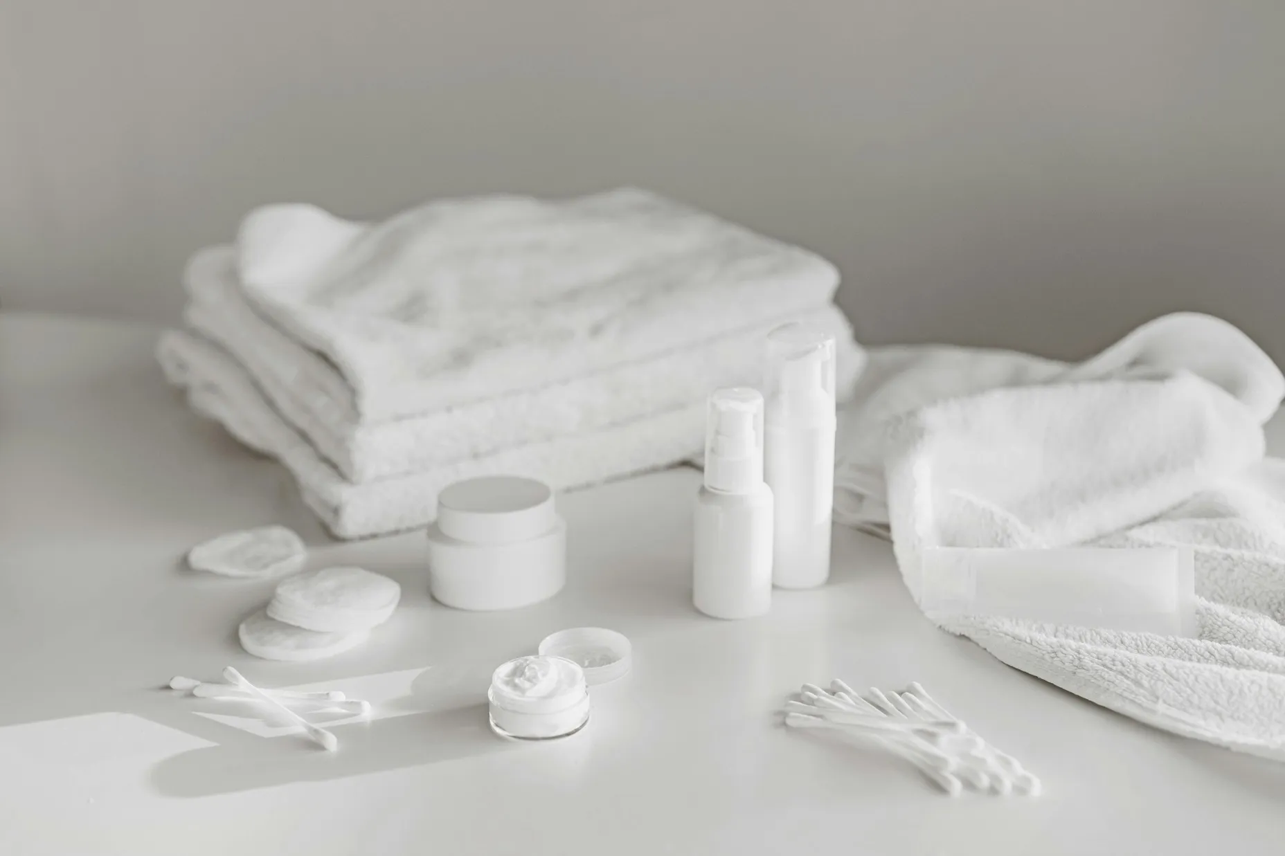 Minimal skincare products with towels, cotton pads, and bottles arranged on a clean white surface in a simple bathroom setting